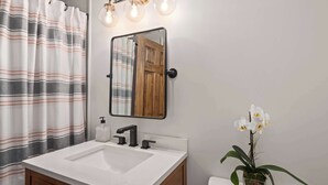 A bathroom with a white sink, wooden cabinets, a mirror with a wooden door reflection, and a potted orchid. - 2BR Stowe Mountain Unit | River & Ski Access (Stowe)