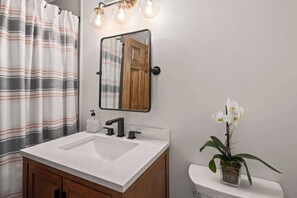 Un baño con lavamanos blanco, armarios de madera, un espejo con reflejo de una puerta de madera y una orquídea en maceta.