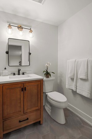 A bathroom with a wooden vanity, white sink, and a toilet. There is a mirror above the sink and a light fixture above it.