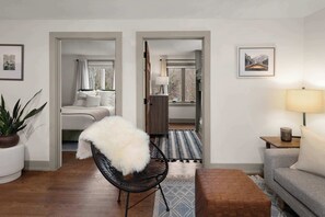 Una sala de estar moderna con un sofá, una butaca, una lámpara y un tapete. Hay una cama en la habitación contigua.