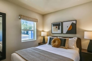 A hotel room with a bed, two bedside lamps, a window with a view, and two framed pictures on the wall. - Deluxe Desert Getaway | Heated Pool & Hot Tub (Scottsdale)