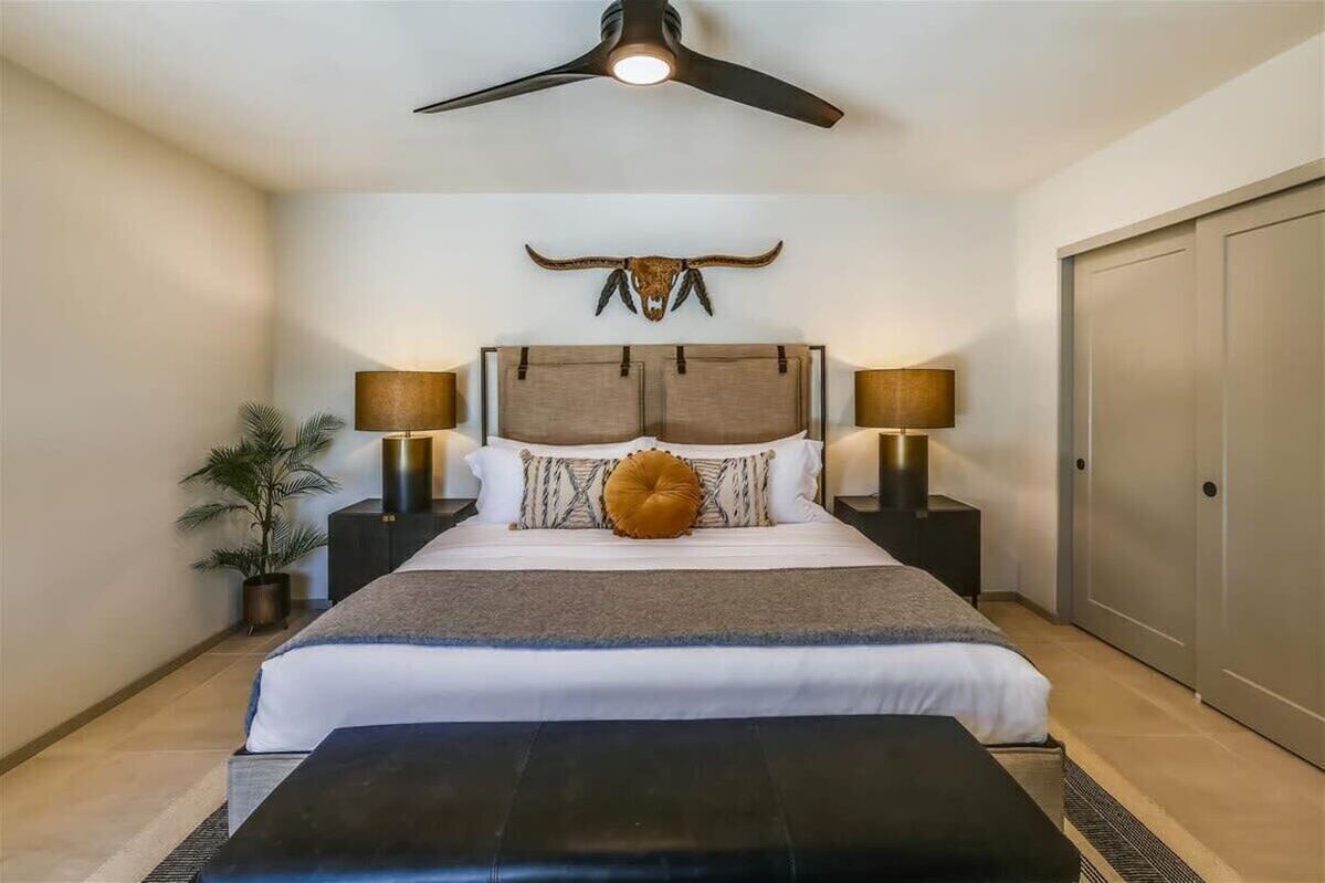 A bedroom with a large bed, two bedside lamps, a ceiling fan, a wall-mounted bull skull decoration, a potted plant, and a sliding door.