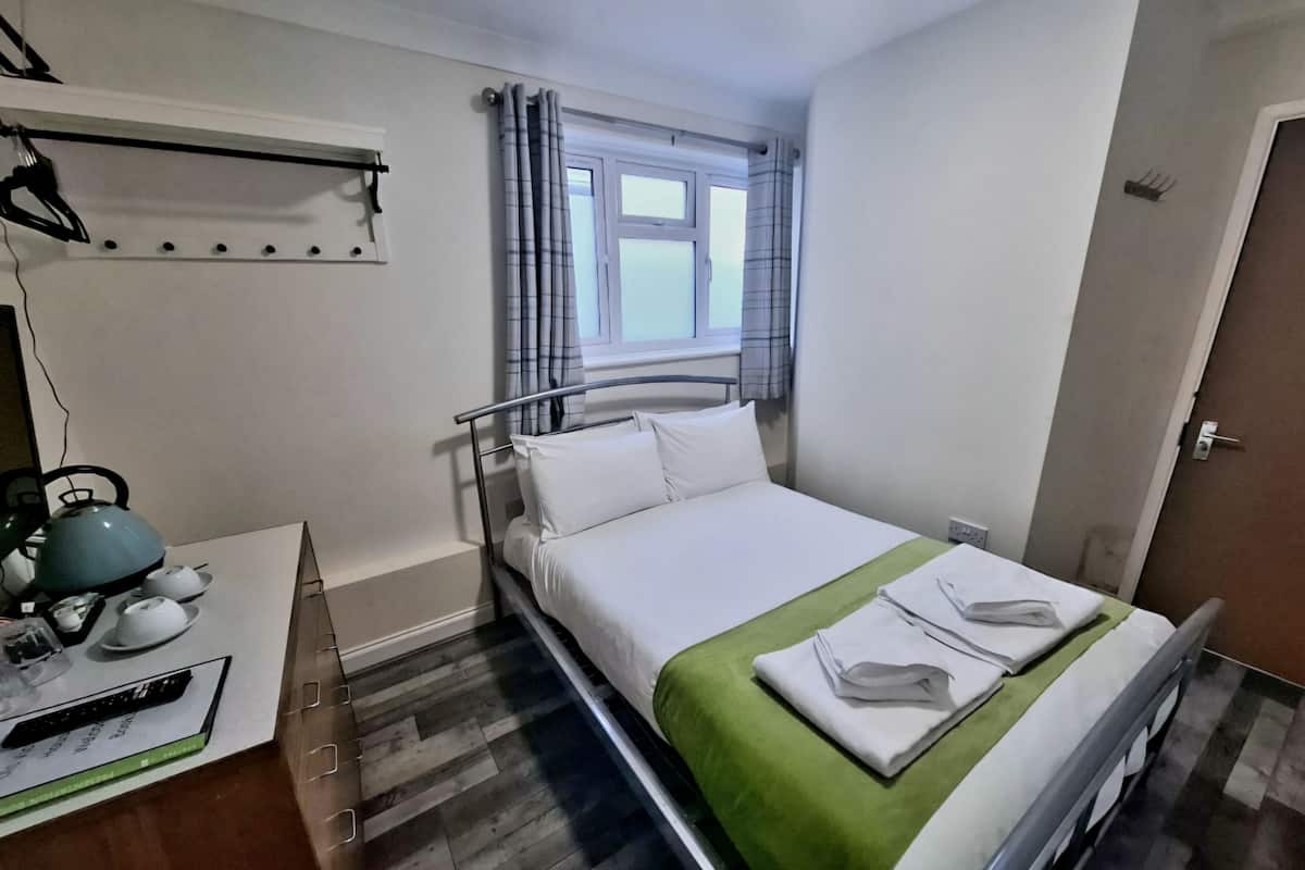 A bedroom with a bed, a window with curtains, a kettle, and a small table with a kettle and cups.