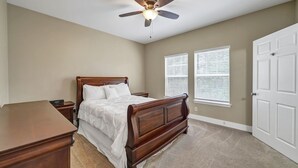 Ein Schlafzimmer mit einem hölzernen Bett, einem Nachttisch, einem Schreibtisch, einem Deckenventilator und zwei Fenster mit Jalousien.