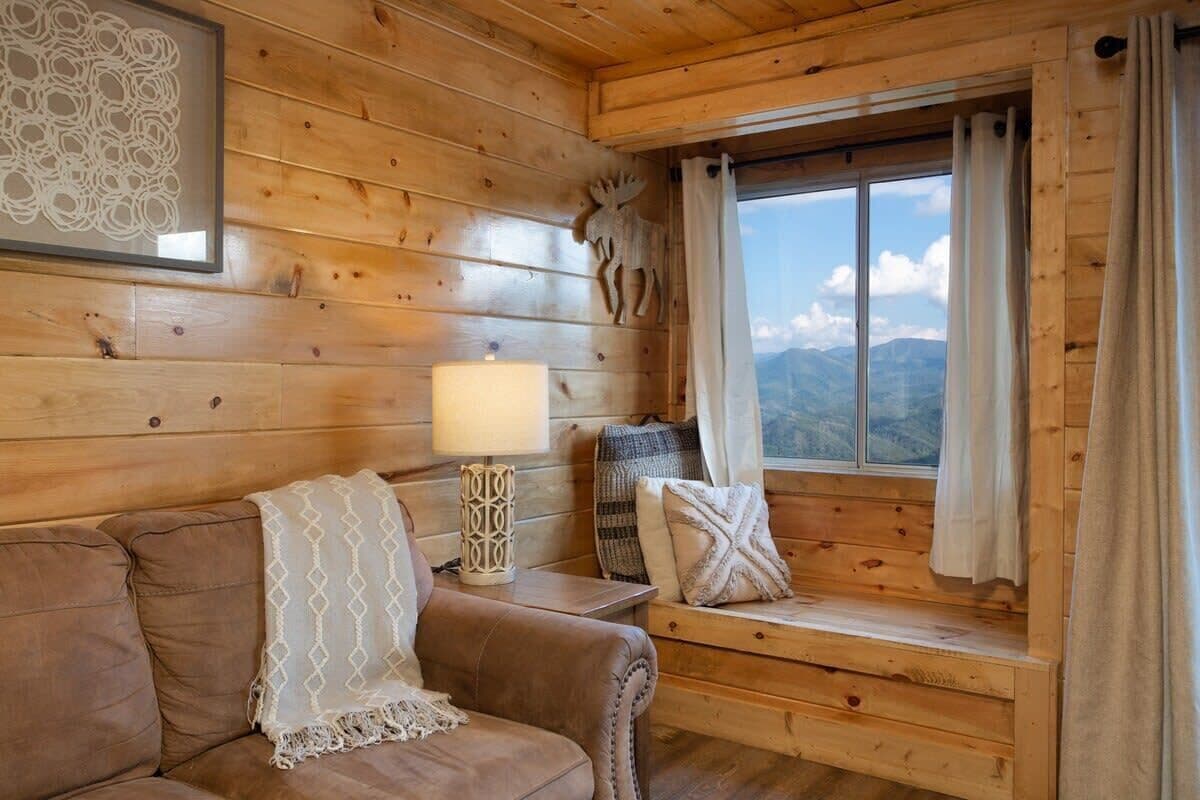 A cozy cabin interior with a wooden wall, a cushioned bench, a lamp, and a window with curtains.