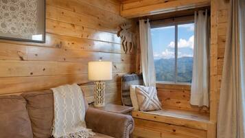 A cozy cabin interior with a wooden wall, a cushioned bench, a lamp, and a window with curtains.
