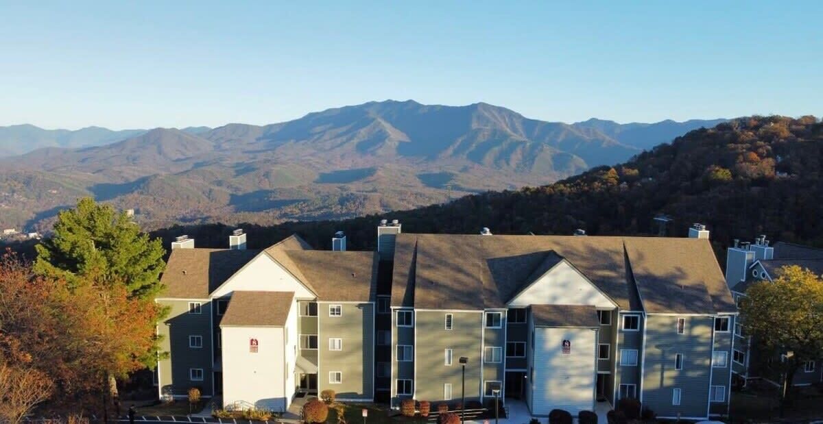 Un edificio de varios pisos con techo plano, rodeado de árboles y montañas al fondo.