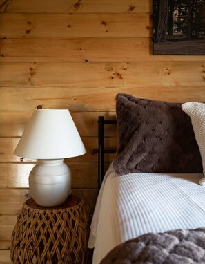 Una pared de madera con una lámpara blanca sobre un taburete tejido, una cama con ropa blanca y una almohada marrón, y un cuadro enmarcado sobre la cama.