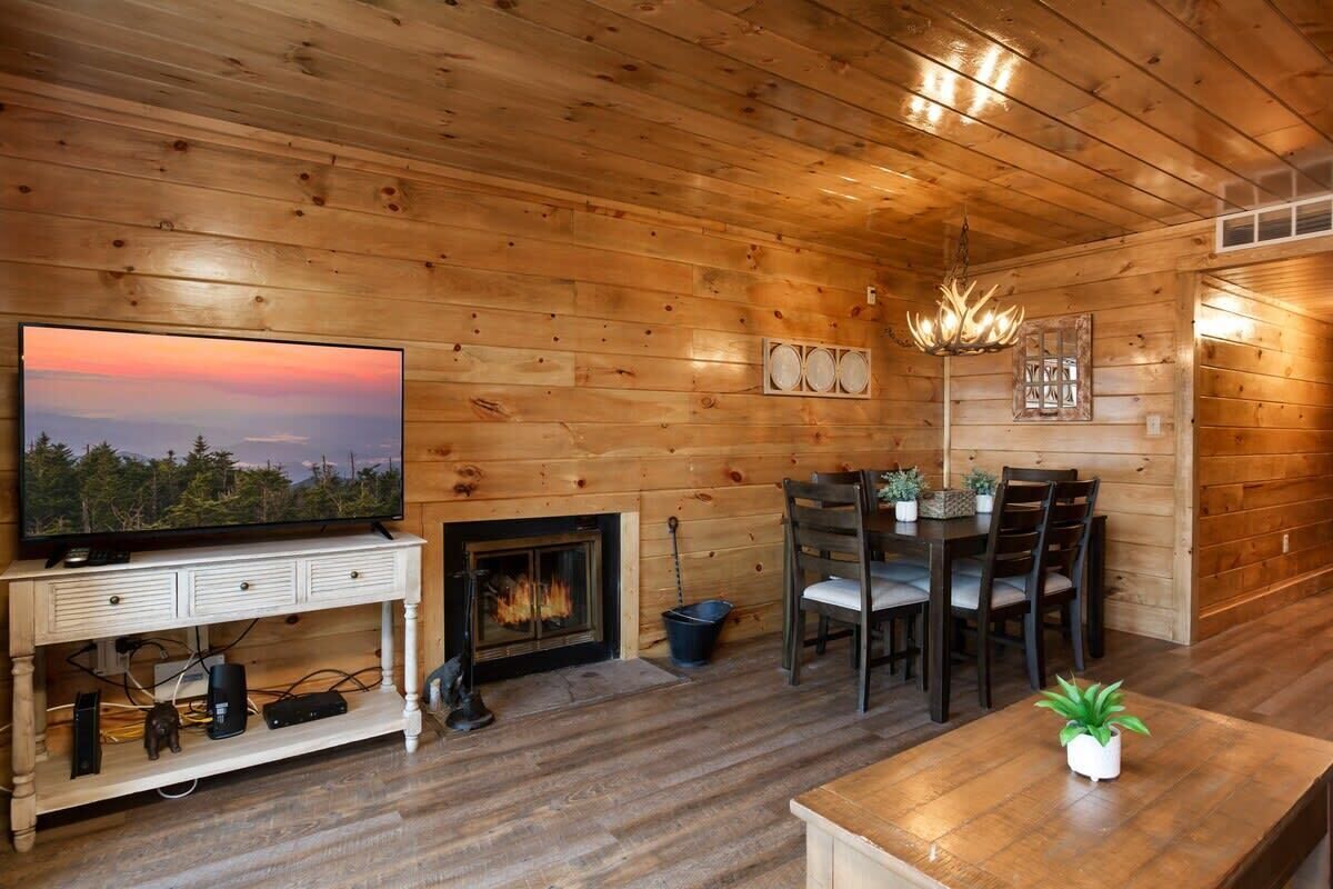 A cozy log cabin interior with a television on a white console, a fireplace, a dining table with chairs, and a chandelier.