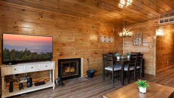 A cozy log cabin interior with a television on a white console, a fireplace, a dining table with chairs, and a chandelier.