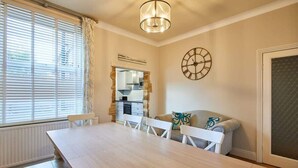 A dining area with a long table, white chairs, a chandelier, a clock, and a view into the kitchen. - Host & Stay | Allium House (Whitby)