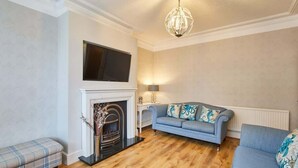 A living room with a fireplace, a blue sofa, a plaid bench, and a television mounted above the fireplace. - Host & Stay | Allium House (Whitby)