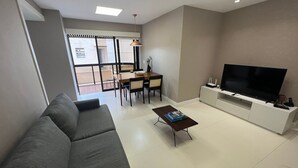 A modern living room with a grey sofa, a dining table with chairs, a coffee table with a book, and a TV on a white entertainment unit. - Ipanema Flat - Vieira Souto 500  (Rio de Janeiro)