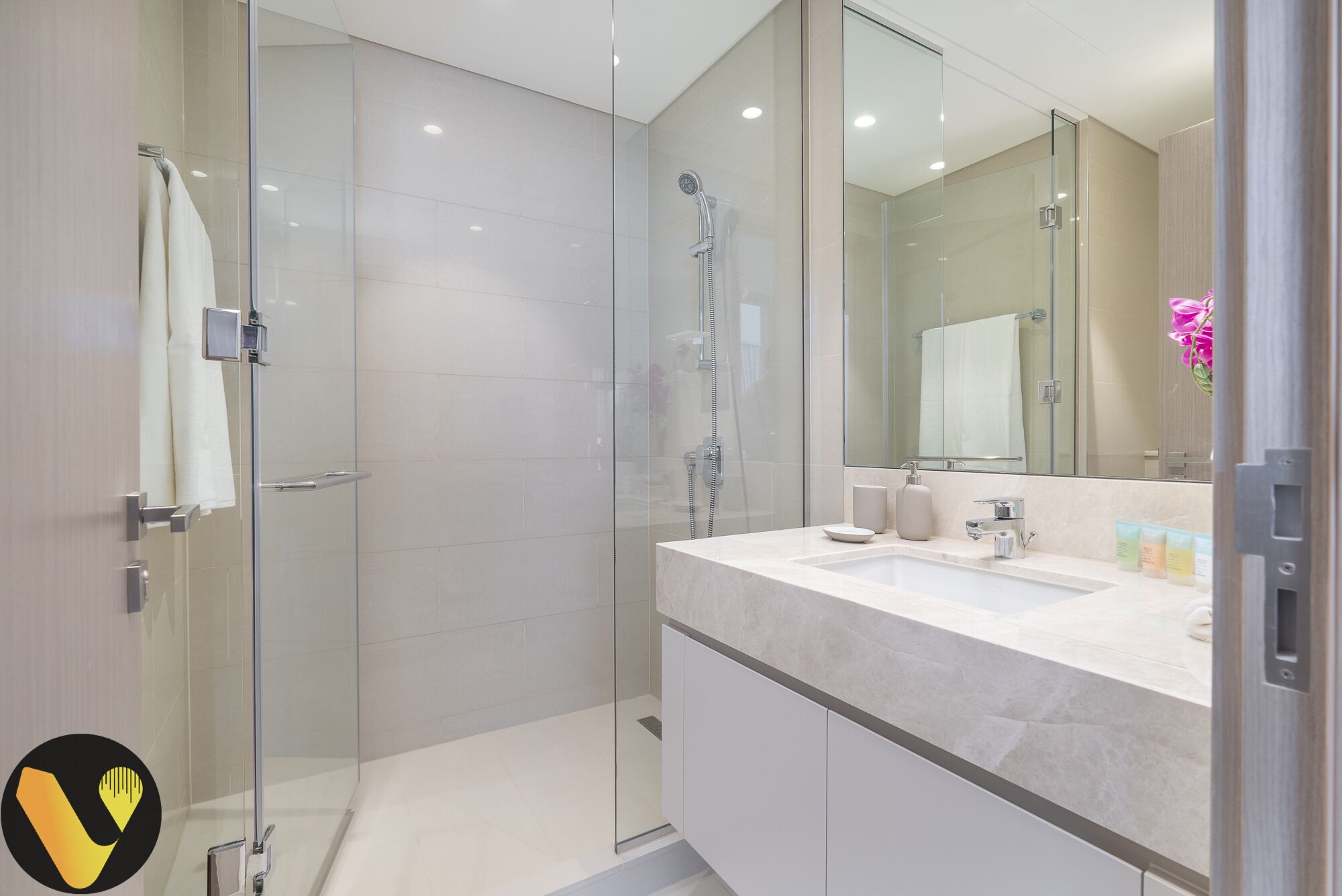 Une salle de bain moderne avec une douche à parois de verre, un grand miroir et un plan de toilette blanc agrémenté d’articles de toilette.