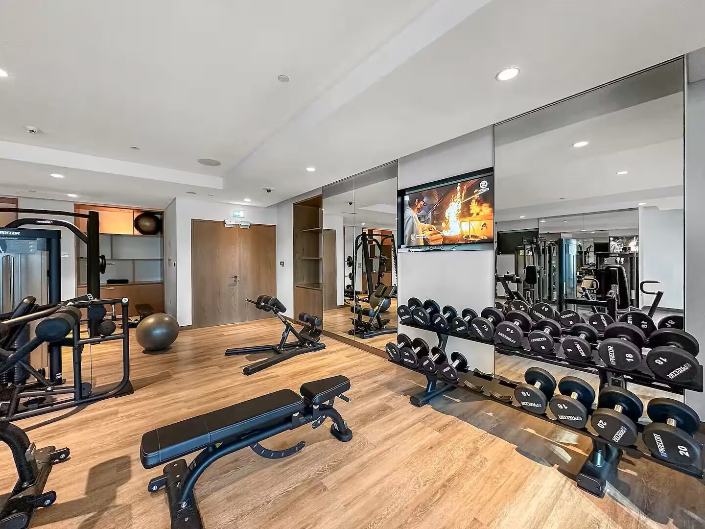 A modern gym with various exercise equipment, a bench, and a wall-mounted TV displaying a workout video.
