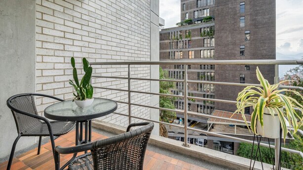 Ein Balkon mit rundem Tisch und Stühlen, Blumentöpfen und Blick auf ein hohes Gebäude mit Balkonen.