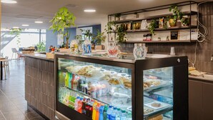 Ein modernes Café mit einer Glastheke, gefüllt mit Gebäck und Getränken. Im Hintergrund sind Regale mit Pflanzen und Küchenutensilien zu sehen.