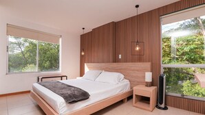 Ein modernes Schlafzimmer mit einem Holzkopfende, einem großen Fenster mit Blick ins Grüne, einem Nachttisch mit Lampe und einem Standheizkörper.