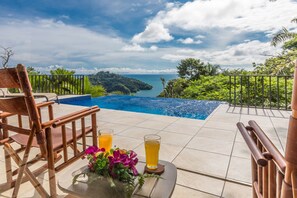 Zona junto a la piscina con vista al océano, una silla y dos bebidas sobre una mesa.