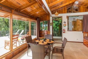 Una terraza de madera con mobiliario de mimbre, una mesa con individuales y vistas al follaje circundante.