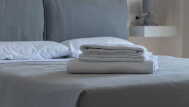 A neatly made bed with white linens and pillows, a folded towel on the bed, and a gray headboard.