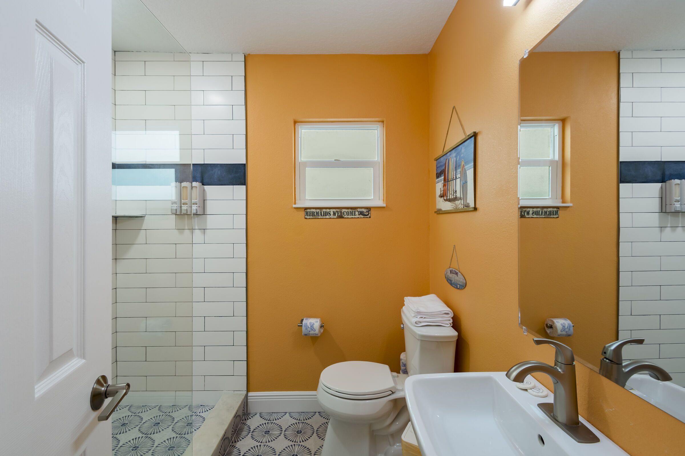 Un baño con inodoro blanco, lavamanos y zona de ducha. Hay dos ventanas y un letrero en la pared.
