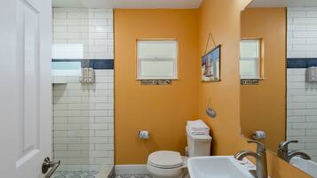 Un baño con inodoro blanco, lavamanos y zona de ducha. Hay dos ventanas y un letrero en la pared.
