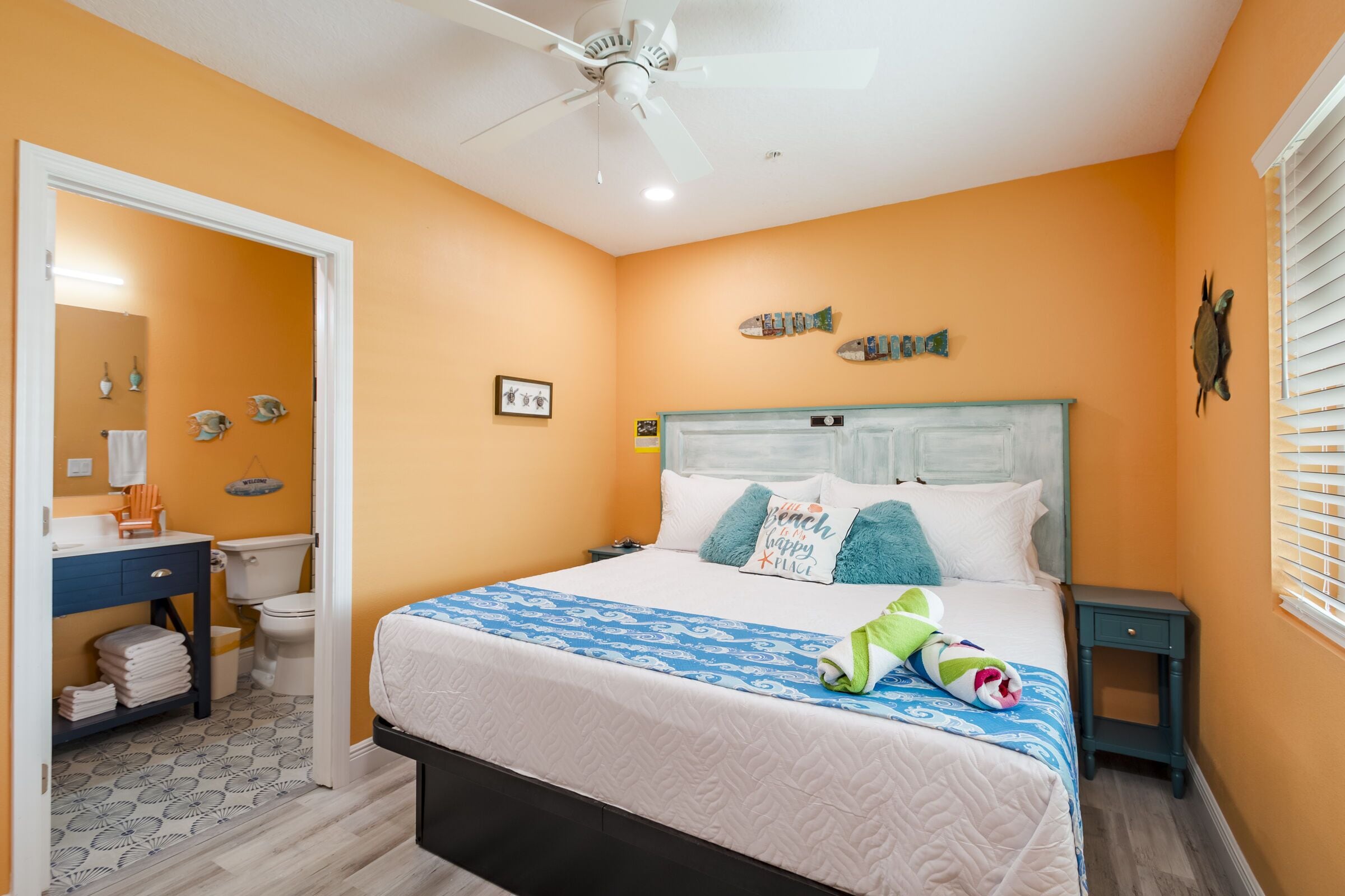 A bedroom with an orange wall, a bed with a blue and white patterned bedspread, a white ceiling fan, and a small nightstand.