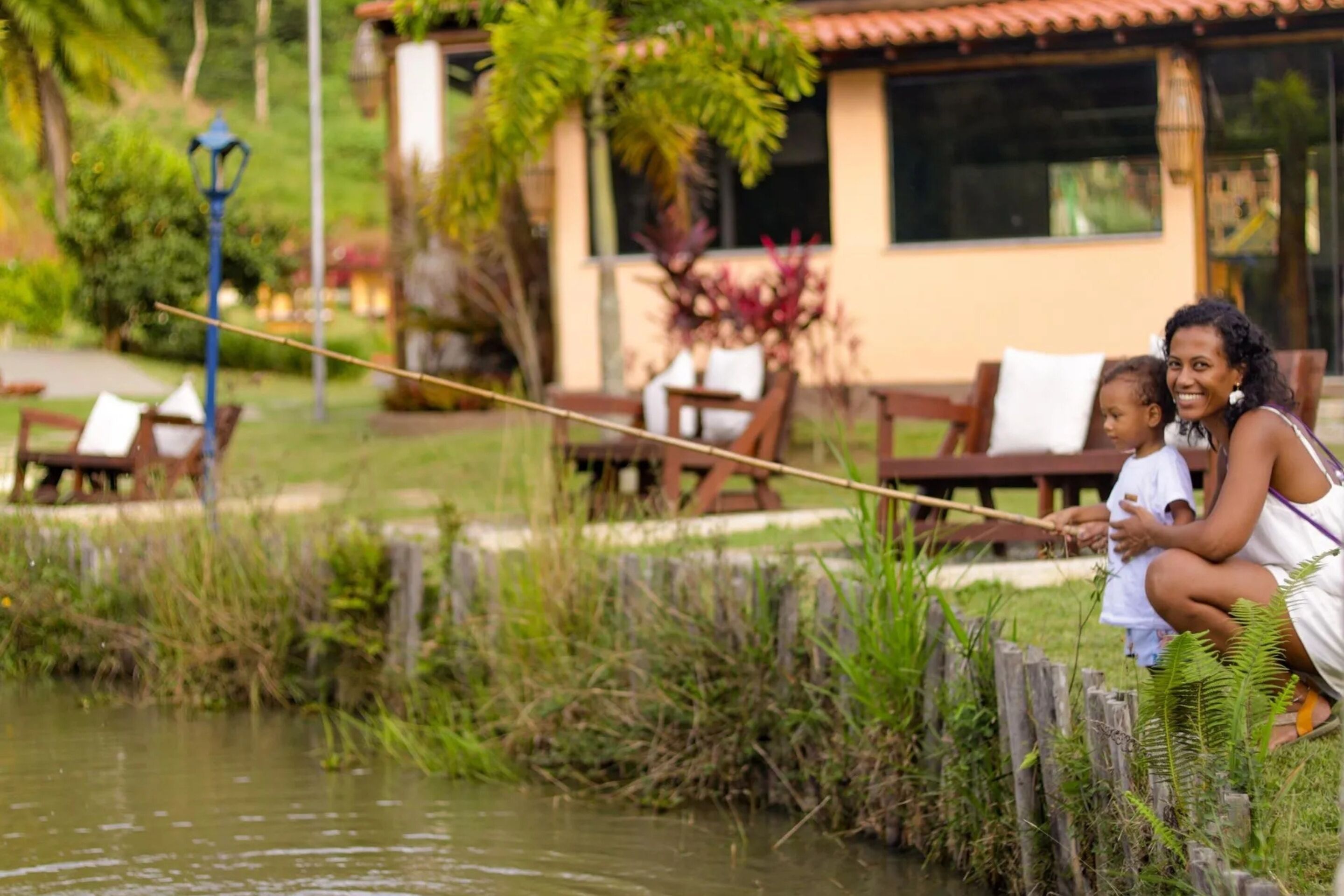 A woman and child fishing by a calm water body, with a wooden fence and greenery in the background.