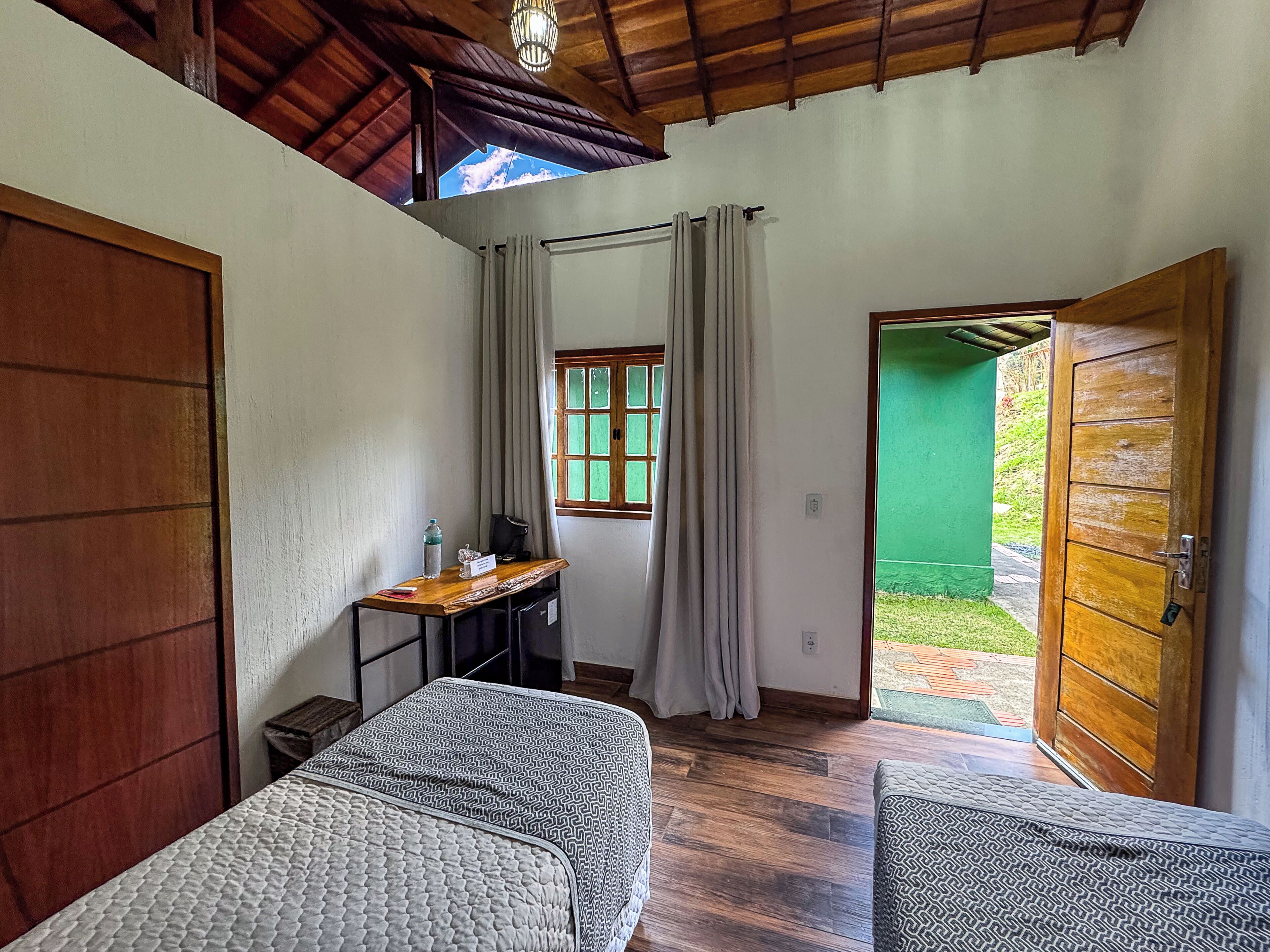 Quarto com cama, escrivaninha, janela com cortinas e porta que dá para o exterior.