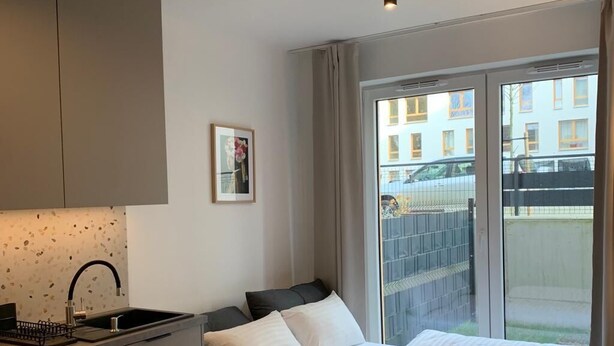 Une chambre d’hôtel compacte, dotée d’une kitchenette, d’un lit avec une literie blanche, d’une petite table ronde et d’une grande fenêtre donnant sur la rue et les bâtiments.