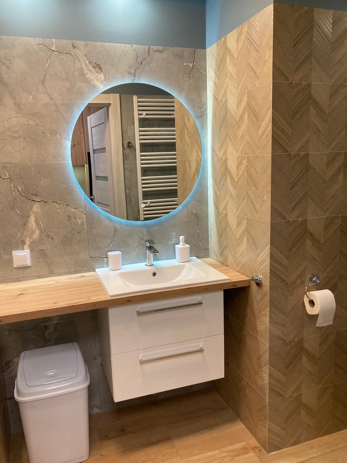 A bathroom with a marble vanity, a round mirror, a wooden countertop, a sink, a toilet paper holder, and a trash bin.