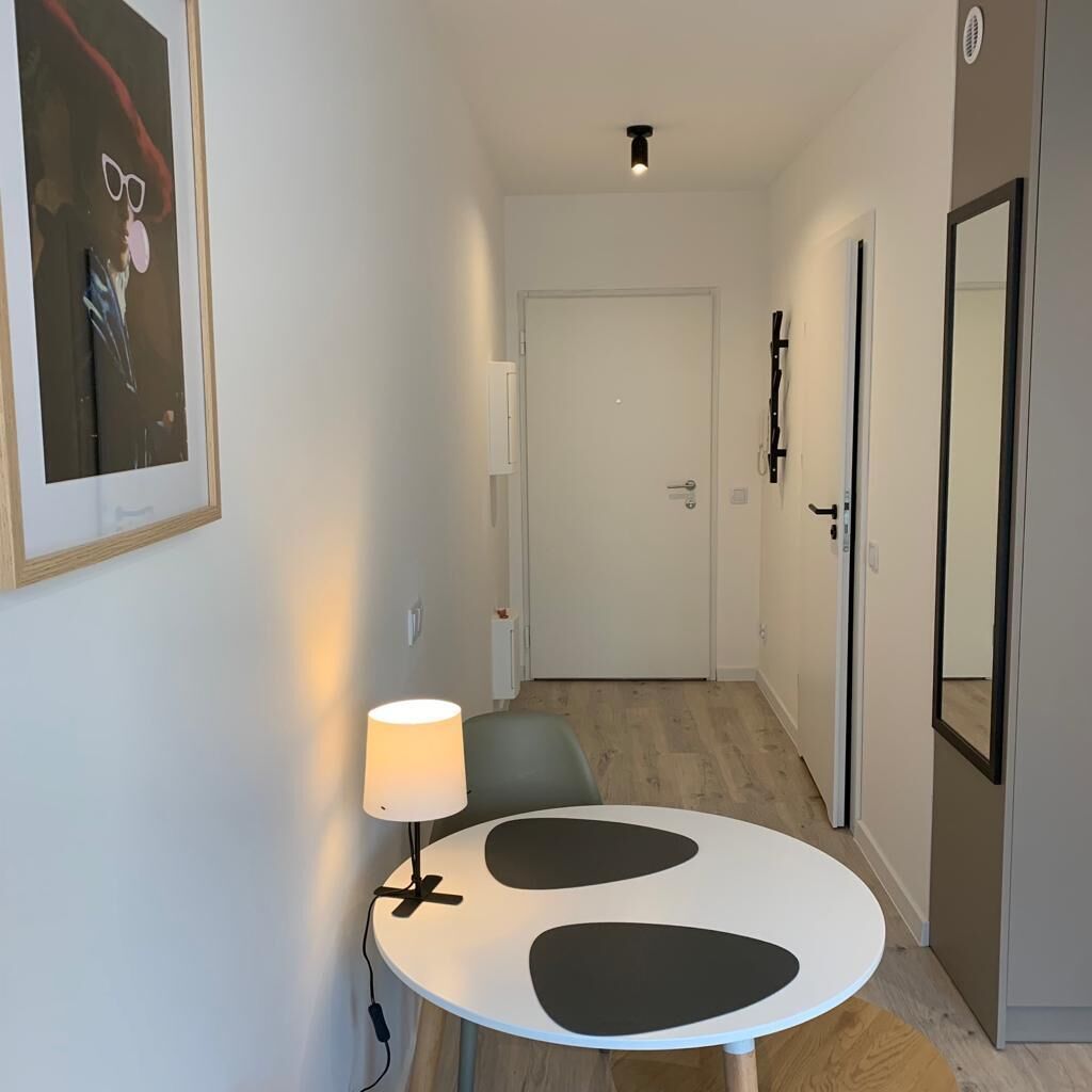 En smal hall med ett runt bord och en lampa, en dörr längst in och en spegel på den högra väggen.