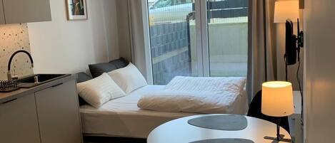 Habitación de hotel compacta con cama, pequeña kitchenette, mesa de comedor con sillas, ventana con cortinas y lámpara fijada a la pared.