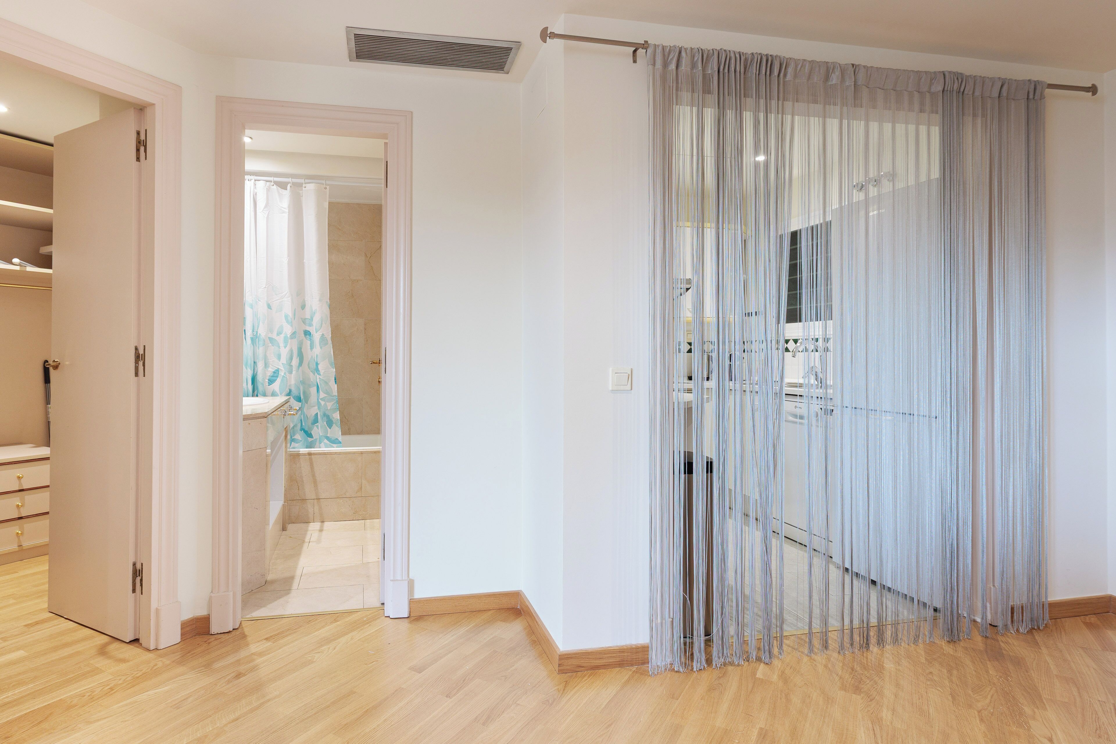 A modern hotel room with a wooden floor, a bathroom with a shower curtain, and a large window with sheer curtains.