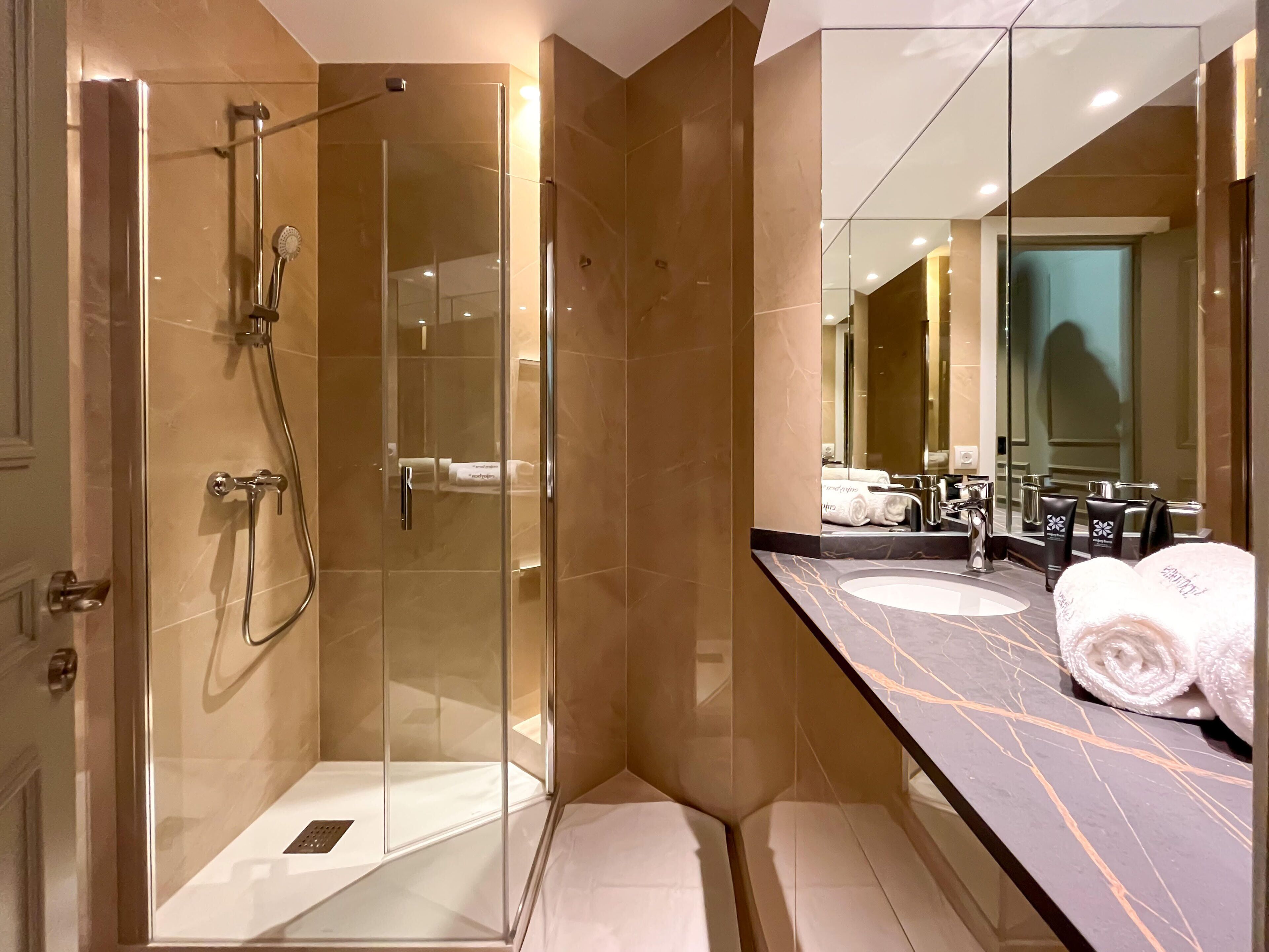 Un baño moderno con ducha envuelta en vidrio, paredes con azulejos de mármol, tocador con lavabo y un espejo grande.