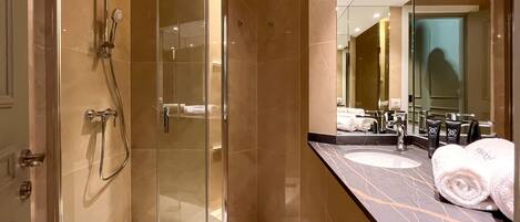 Un baño moderno con ducha envuelta en vidrio, paredes con azulejos de mármol, tocador con lavabo y un espejo grande.