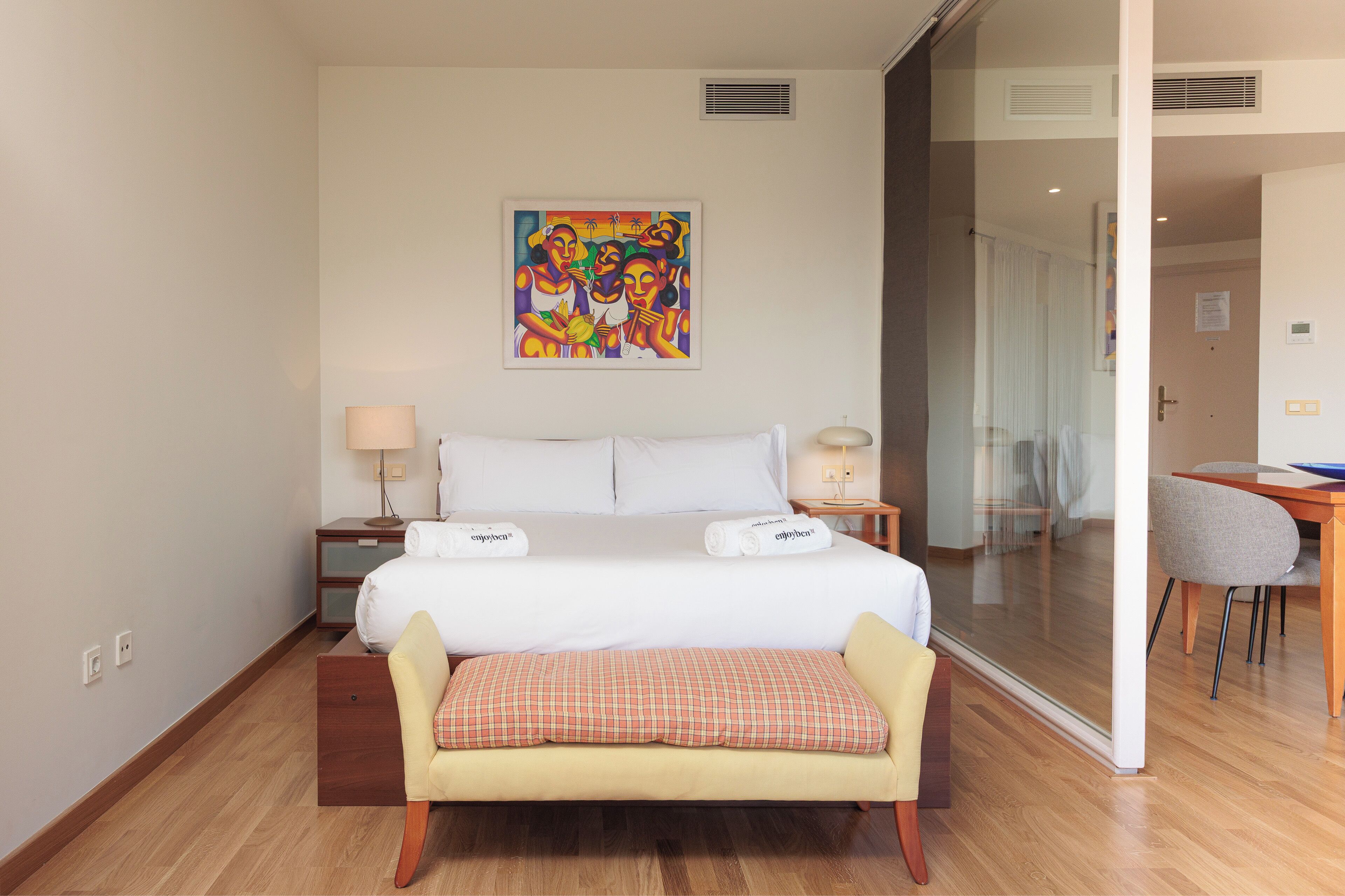 A hotel room with a bed, bedside tables, a bench, a painting, and a view through a glass door.