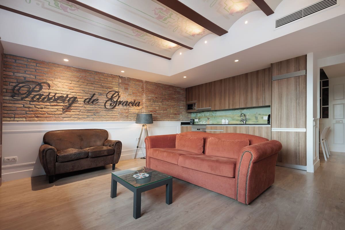 Una sala moderna con una pared de ladrillo decorativa, un sofá marrón, una mesa de centro de vidrio y una zona de cocina con armarios de madera y un microondas.