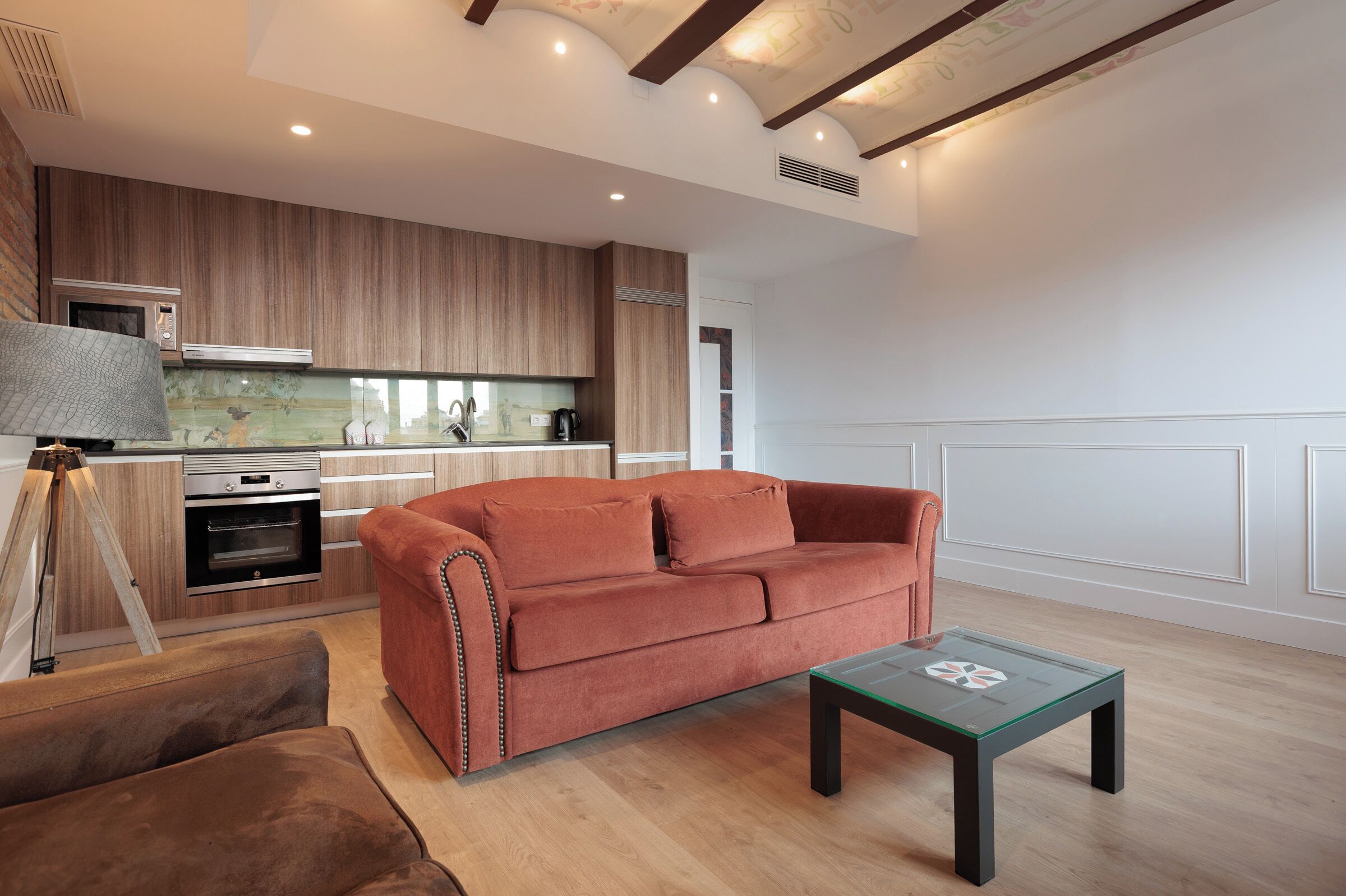 En moderne stue med sofa, sofabord og et køkkenområde med træskabe og hårde hvidevarer.