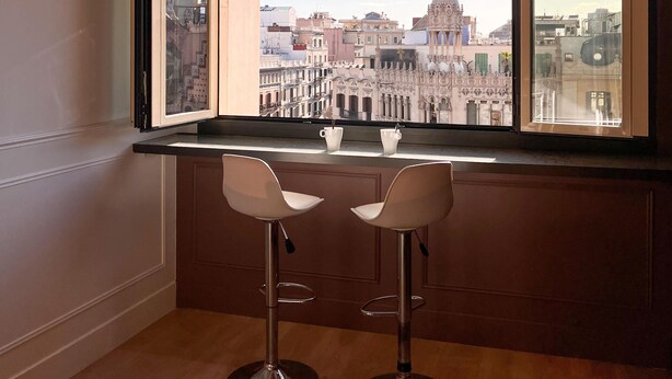 Una habitación con un amplio ventanal que da a un paisaje urbano, dos taburetes de bar y un techo decorativo.