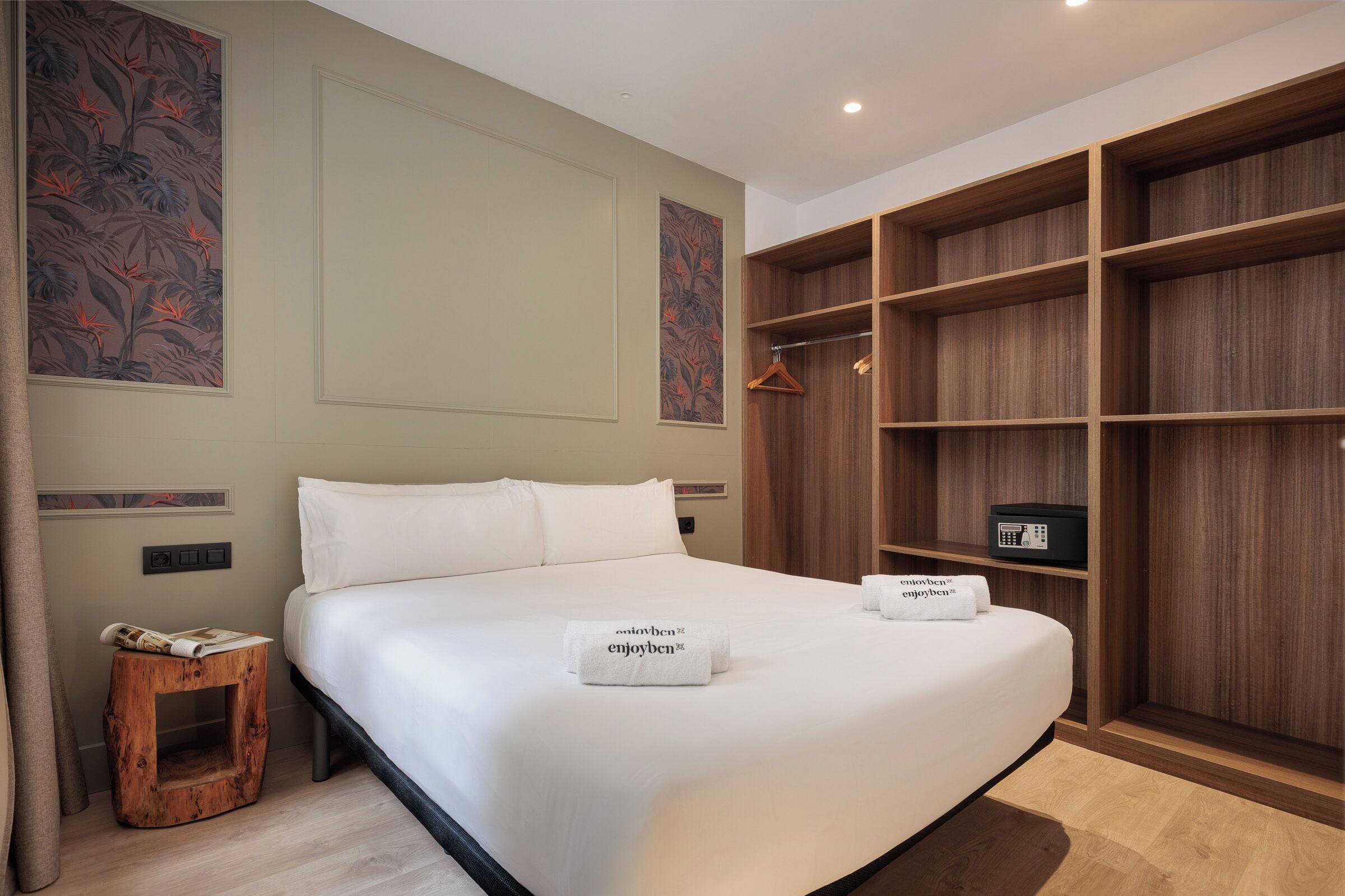 Una cama bien hecha con dos toallas, una mesita de madera, un armario de madera y un taburete pequeño de madera.