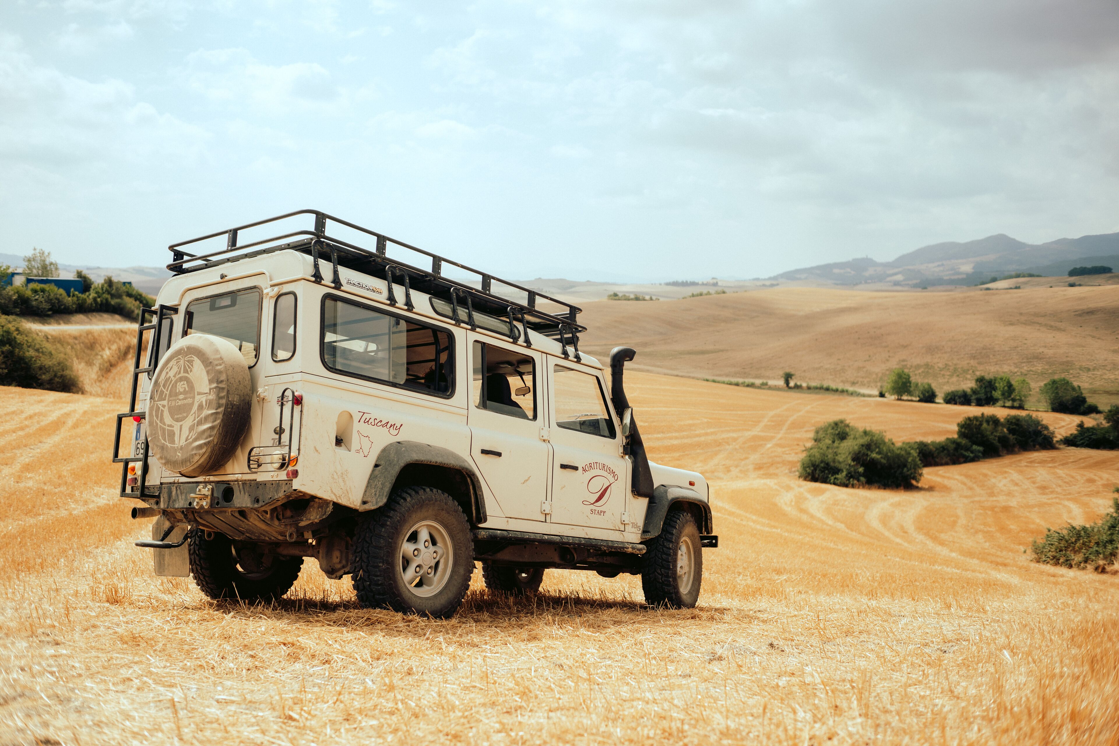 A white SUV with a spare tire mounted on the back, parked on a dry, golden field with rolling hills in the background.