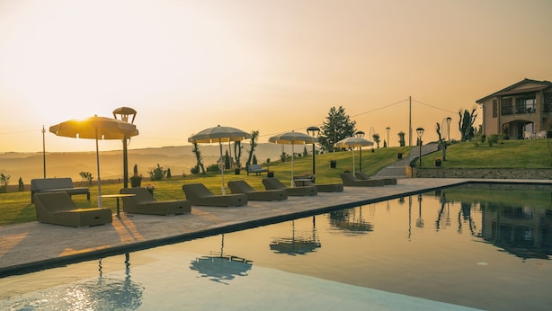 A poolside area with lounge chairs and umbrellas, a clear view of the pool, and a building in the background. - Villa Etru (Peccioli)
