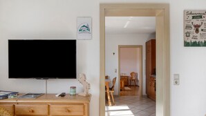 Una stanza con televisione, arredi in legno, un'area pranzo con sedie e tavolo, e una cucina visibile attraverso una porta aperta.