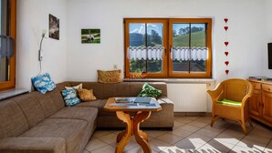 Un soggiorno con un divano marrone, un tavolino in legno, una poltrona in vimini, una finestra vista su un paesaggio verde e un termosifone.