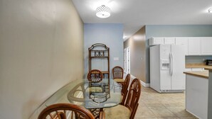 A dining area with a glass table and wooden chairs, a white refrigerator, and a kitchen area with white cabinets. - 30A Getaway Too! (Santa Rosa Beach)