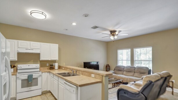 A modern kitchen with white cabinets, a stove, and a dishwasher. There is a dining area with a sofa and a TV. The room has large windows and a ceiling fan. - 30A Getaway Too! (Santa Rosa Beach)