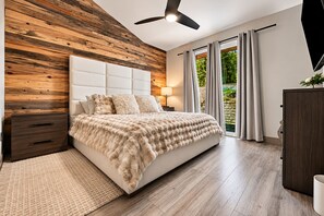 Un dormitorio con una pared decorativa de madera, un cabecero capitonado, una cama grande con una manta mullida, una mesita de noche, una lámpara y vistas a la vegetación por la ventana.