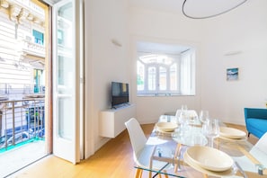 A modern dining area with a glass table, white chairs, and a TV on a white cabinet.